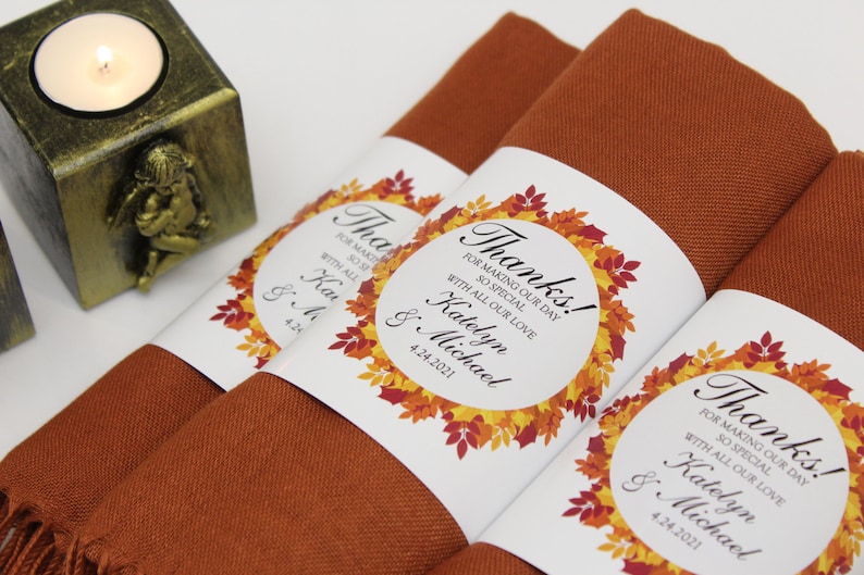 May include: Close-up of folded rust-colored napkins with white labels. The labels feature a fall leaf wreath design and the text "Thanks!" along with names and a date. A gold candle holder with an angel is in the background.