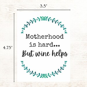 May include: A white card with a green leafy wreath design around the text "Motherhood is hard... But wine helps". The card measures 3.5 inches by 4.75 inches.