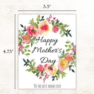 May include: A white card with a floral wreath design in shades of pink, yellow, and green. The text "Happy Mother's Day" is in the center of the wreath, and the text "To the best mom ever" is at the bottom of the card. The card measures 4.75 inches by 3.5 inches.
