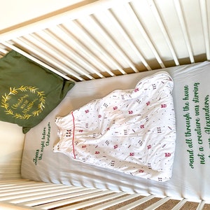 May include: A white cot with a green pillow that says "Christmas with the Matthews" and a white sleeping bag with red and black accents. The cot sheet is grey with green text that says "Twas the night before Christmas and all through the house not a creature was stirring not even Alexander."