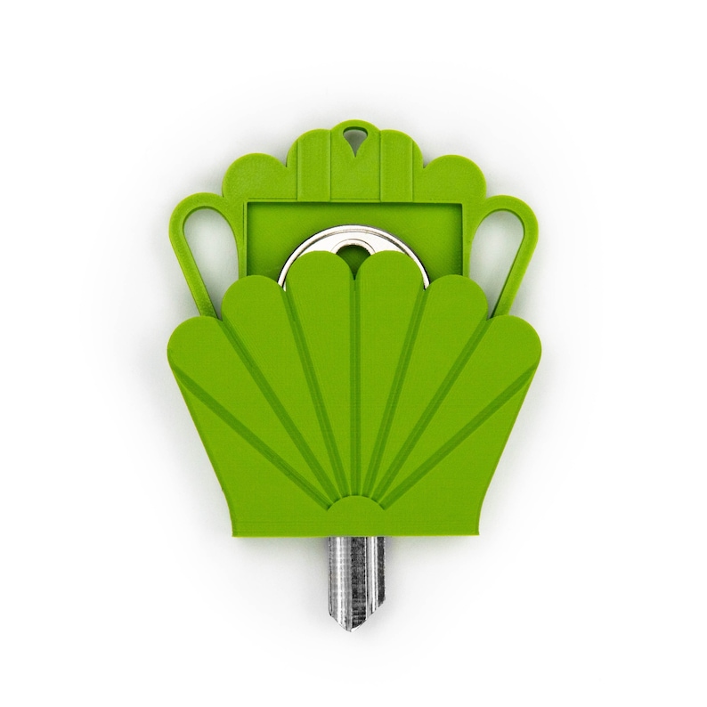 May include: A bright lime green plastic key cover shaped like a stylized shell or fan with a small hole at the top for a keychain. The cover features a decorative scalloped edge and two side handles, partially enclosing a silver metal key blade.