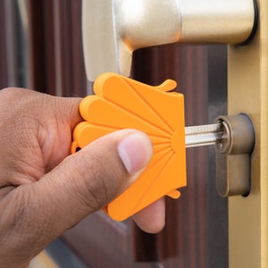 May include: A bright orange plastic key turner with a fan-shaped grip featuring raised ridges. The device is attached to a metal key inserted into a door lock cylinder, designed to provide extra leverage for turning the key in the lock mechanism.