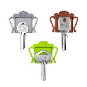 May include: Three metal keys with JMA branding, each inserted into a decorative plastic key cap. The caps are shaped like trophies with handles and come in three distinct colors: a textured silver-gray, a textured brown, and a smooth lime green.