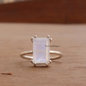 Puede incluir: Un anillo de plata con una piedra lunar rectangular e iridiscente. La piedra lunar está engastada con cuatro garras. La banda del anillo es delgada y lisa. El anillo se muestra sobre una superficie de madera. El texto "TheSilverEmpire" es visible.