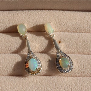 Puede incluir: Un par de pendientes colgantes de plata con piedras de ópalo y un halo de cristal transparente. Los pendientes tienen forma de lágrima.