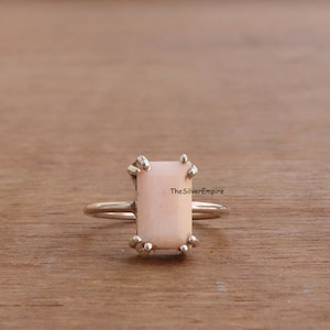 May include: A silver ring with a rectangular pink opal gemstone set in a prong setting. The ring is on a wooden surface. TheSilverEmpire is printed on the image.