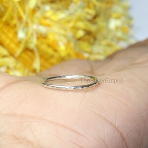 May include: A thin silver ring with a hammered texture. The ring is on a hand, with a blurred yellow background.