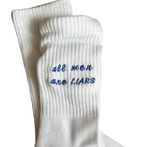 Könnte beinhalten: Weiße Socken mit dem gestickten Text "all men are LIARS" in Blau. Die Socken sind am oberen Rand gerippt und haben die Größenangabe "39-46" unten aufgedruckt. Die Socken sind aus weichem Material.