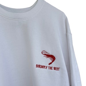 Puede incluir: Sudadera blanca con un camarón bordado en rojo y el texto "Shrimply the Best". La sudadera tiene cuello redondo y mangas largas. El diseño está en la parte superior izquierda del pecho.