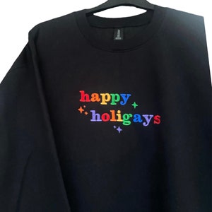 Könnte beinhalten: Schwarzes Sweatshirt mit dem gestickten Text "happy holigays" in Regenbogenfarben. Das Design beinhaltet Sternakzente. Das Sweatshirt hängt an einem schwarzen Kleiderbügel.