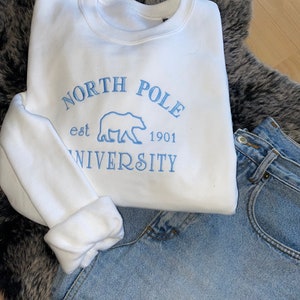 May include: A white sweatshirt with the embroidered text "NORTH POLE UNIVERSITY est 1901" in light blue, along with a polar bear graphic. The sweatshirt is paired with light-wash denim jeans, resting on a gray faux fur rug.