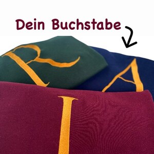 Può includere: Primo piano di tre tessuti ricamati, ognuno con una lettera dorata. I tessuti sono nei colori bordeaux, verde scuro e blu navy. Il testo tedesco "Dein Buchstabe" è in alto.