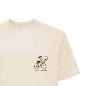 Könnte beinhalten: Cremefarbenes T-Shirt mit einer kleinen braunen Illustration einer Person, die Fahrrad fährt, mit Dampf, der aus einer Kaffeetasse aufsteigt. Der Text "- cycling for coffee-" steht darunter. Das Shirt hat einen Rundhalsausschnitt und kurze Ärmel.