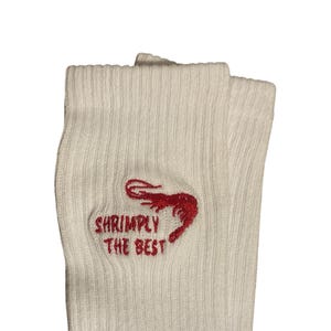 May include: A pair of white ribbed socks with the embroidered text "Shrimply The Best" and a red shrimp graphic. The socks are folded over, showcasing the design on the front.