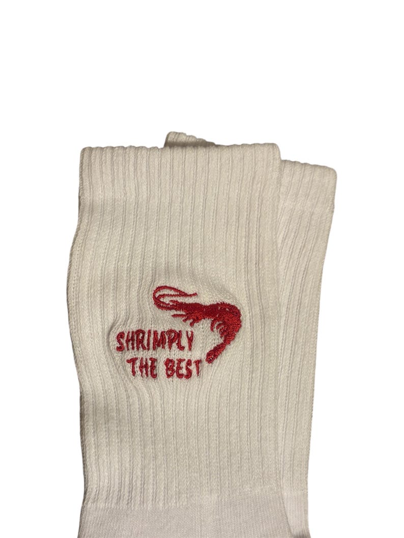 May include: A pair of white ribbed socks with the embroidered text "Shrimply The Best" and a red shrimp graphic. The socks are folded over, showcasing the design on the front.