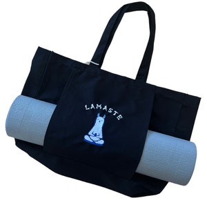 Op de afbeelding: Zwarte canvas draagtas met een lichtgrijze yogamat. De tas heeft een zak met het geborduurde woord "LAMASTE" en een mediterend lama-ontwerp in wit en blauw. De tas heeft twee handvatten.