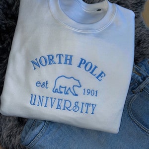 May include: A white sweatshirt with blue embroidered text that reads "NORTH POLE UNIVERSITY est 1901" with a bear graphic. The sweatshirt is paired with blue jeans, resting on a gray faux fur surface.