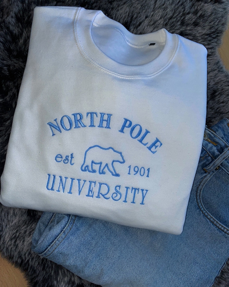 May include: A white sweatshirt with blue embroidered text that reads "NORTH POLE UNIVERSITY est 1901" with a bear graphic. The sweatshirt is paired with blue jeans, resting on a gray faux fur surface.