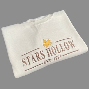 May include: Cream-colored sweatshirt with the embroidered text "STARS HOLLOW" and "EST. 1779" in brown, along with a yellow leaf graphic. The sweatshirt is a casual, crew-neck style.