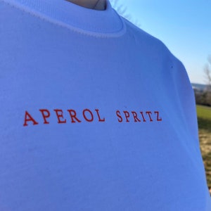 May include: White crew neck t-shirt with the words "APEROL SPRITZ" printed in orange across the chest. The shirt is made of a soft material and is being worn outdoors on a sunny day.