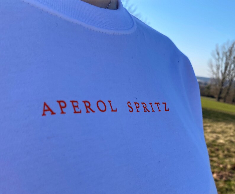 May include: White crew neck t-shirt with the words "APEROL SPRITZ" printed in orange across the chest. The shirt is made of a soft material and is being worn outdoors on a sunny day.