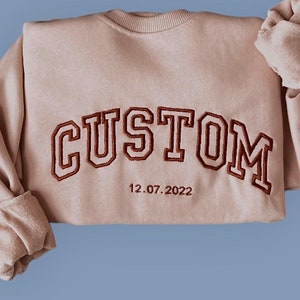 Könnte beinhalten: Beiges Sweatshirt mit dem Wort "CUSTOM" in braunen Blockbuchstaben gestickt. Unter dem Wort steht das Datum "12.07.2022". Die Ärmel sind umgeschlagen, und der Stoff wirkt weich.