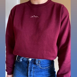 Könnte beinhalten: Ein weinrotes Sweatshirt mit einem kleinen, weiß gestickten Bergketten-Design auf der Brust. Die Person trägt blaue Jeans. Das Sweatshirt hat lange Ärmel und einen Rundhalsausschnitt.
