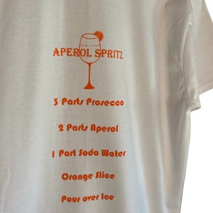 May include: White t-shirt featuring an orange Aperol Spritz cocktail recipe. The recipe includes Prosecco, Aperol, soda water, and an orange slice. The text is printed in orange.