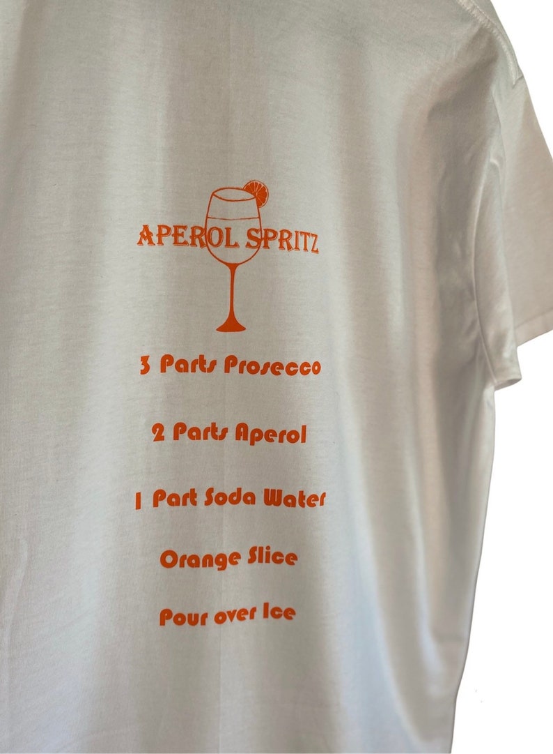 May include: White t-shirt featuring an orange Aperol Spritz cocktail recipe. The recipe includes Prosecco, Aperol, soda water, and an orange slice. The text is printed in orange.