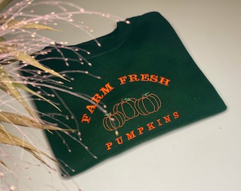 May include: A dark green sweatshirt with the words "FARM FRESH PUMPKINS" embroidered in orange. The design includes three orange pumpkins. The sweatshirt is a fall-themed apparel item.