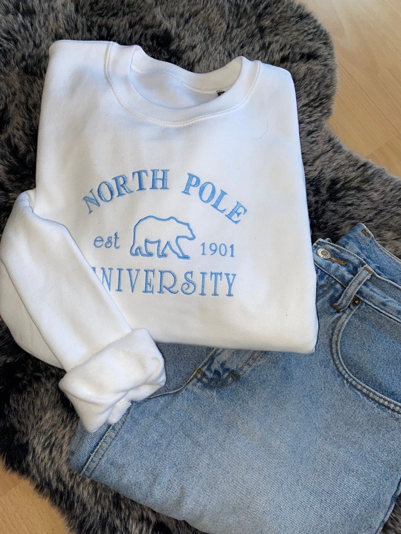 May include: A white sweatshirt with the embroidered text "NORTH POLE UNIVERSITY est 1901" in light blue, along with a polar bear graphic. The sweatshirt is paired with light-wash denim jeans, resting on a gray faux fur rug.