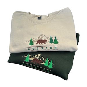 Puede incluir: Dos sudaderas apiladas, una beige y otra verde oscuro. Cada sudadera presenta un diseño bordado de una cordillera, un oso y pinos, con la palabra "ROCKIES" debajo. El diseño es verde, marrón y blanco.