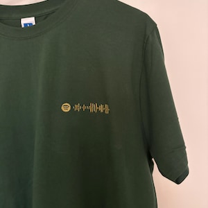 Puede incluir: Camiseta verde oscuro con un pequeño código Spotify dorado bordado en el pecho. La camiseta tiene cuello redondo y mangas cortas. La tela parece suave y el diseño es adecuado para el uso diario. Estilo sencillo e informal.