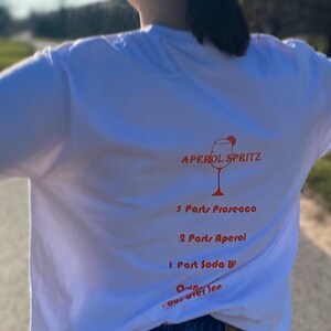 May include: White t-shirt with an Aperol Spritz recipe printed on the back. The recipe includes Prosecco, Aperol, Soda Water, and ice. The design features a graphic of a cocktail glass.