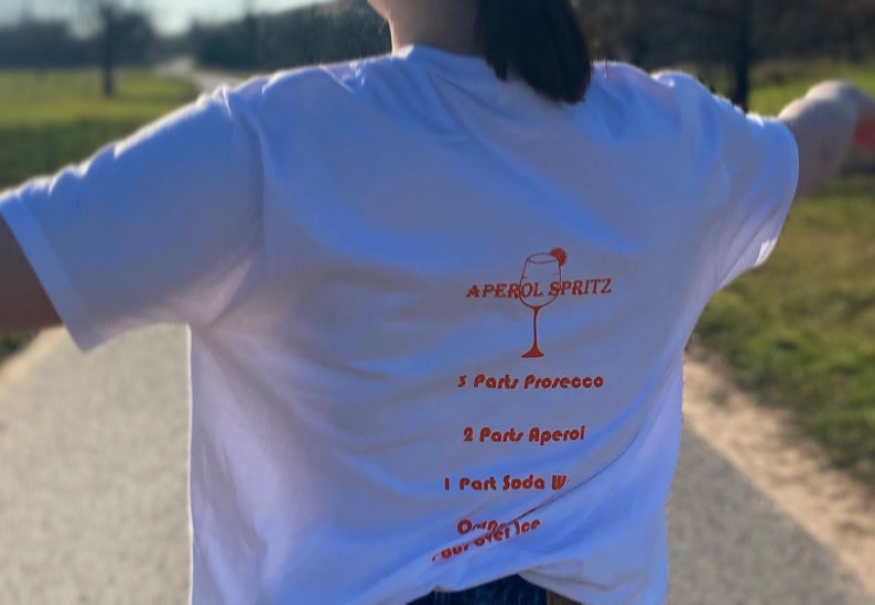 May include: White t-shirt with an Aperol Spritz recipe printed on the back. The recipe includes Prosecco, Aperol, Soda Water, and ice. The design features a graphic of a cocktail glass.