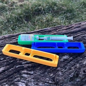 Może przedstawiać: Trzy 3D-wydrukowane plastikowe uchwyty do strzykawki. Uchwyty są żółte, niebieskie i zielone. Strzykawka znajduje się w zielonym uchwycie. Uchwyty znajdują się na drewnianej powierzchni.