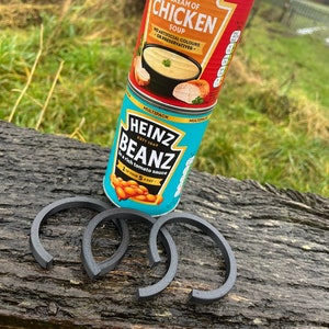 Può includere: Due lattine di zuppa Heinz su una superficie di legno. La lattina superiore è rossa e dice "Heinz Cream of Chicken Soup". La lattina inferiore è blu e dice "Heinz Beanz in a rich tomato sauce". Ci sono quattro anelli di gomma neri sulla superficie.