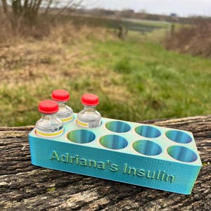 Puede incluir: Un organizador de almacenamiento de insulina impreso en 3D de color azul turquesa con 8 compartimentos. El organizador tiene el texto "Adriana's Insulin" impreso en la parte delantera. Tres pequeños viales de vidrio con tapas rojas están en el organizador.