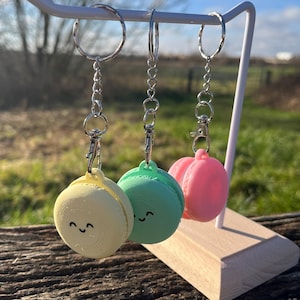 Puede incluir: Tres llaveros de macarons coloridos con caras felices. Los llaveros son amarillos, verdes y rosas y cuelgan de llaveros plateados.