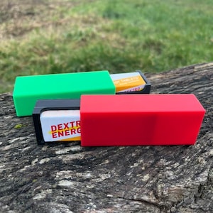 May include: Three rectangular cases, one green and one red, partially open to reveal white and yellow packaging with the text "DEXTRO ENERGY". The cases are resting on a weathered, brown log, with a blurred green background.