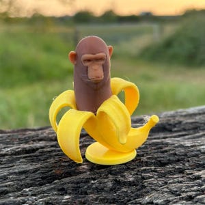 Puede incluir: Una figura caprichosa de un mono saliendo de un plátano pelado. El mono tiene la cara y el cuerpo marrones, con una cáscara de plátano amarilla. La figura está sobre un fondo natural borroso.