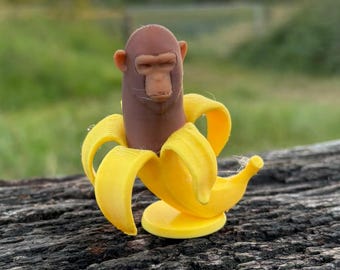 May include: A novelty monkey figurine emerging from a yellow banana peel. The monkey has a brown face and body. The figurine is set against a blurred natural background of green grass and a sunset.