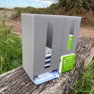 Könnte beinhalten: Ein grauer, 3D-gedruckter Kunststoff-Organizer mit vertikalen Schlitzen für medizinischen Bedarf. Die Einheit enthält mehrere weiße und grüne Dexcom G7 Sensorboxen sowie eine blau-weiße medizinische Applikatorverpackung an der Unterseite.