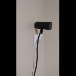 May include: A black cylindrical electronic sensor mounted on a white wall-mounted bracket. The sensor is positioned horizontally and features a black cable extending downwards from its base, secured by the white support structure.