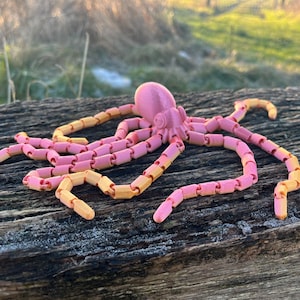 May include: A pink and yellow articulated octopus toy made of plastic. The octopus is posed on a wooden surface.