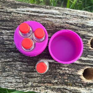 Peut inclure: Un r&eacute;cipient rond violet avec trois petits flacons en verre avec des bouchons rouges. Le r&eacute;cipient est ouvert et les flacons sont visibles. Le r&eacute;cipient est pos&eacute; sur une surface en bois.