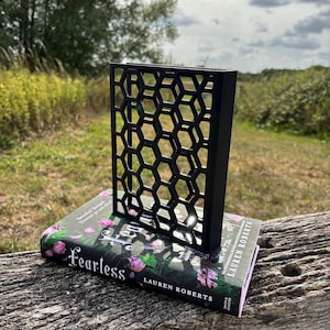 May include: A black metal book stand with a honeycomb design is displayed on top of a book titled "Fearless" by Lauren Roberts. The book has a floral design with pink and purple flowers. The background is a natural outdoor setting.