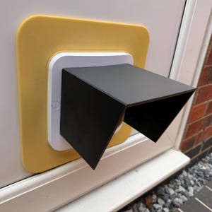 Może przedstawiać: Uchwyt do kamery bezpieczeństwa z kwadratową żółtą płytką tylną, białą kwadratową podstawą i czarną prostokątną osłoną wystającą na zewnątrz. Urządzenie jest zamontowane na białej powierzchni, zapewniając osłonę dla obiektywu kamery.