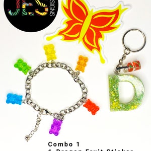 May include: A combo set of three items: a dragon fruit sticker, a glitter letter keychain, and a bracelet with rainbow gummy bear charms. The bracelet is silver and has a lobster clasp.
