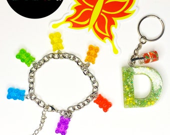 Peut inclure: Un ensemble de trois articles : un autocollant de fruit du dragon, un porte-clés en forme de lettre pailletée et un bracelet avec des breloques en forme d’ours en gomme arc-en-ciel. Le bracelet est argenté et possède un fermoir à ressort.